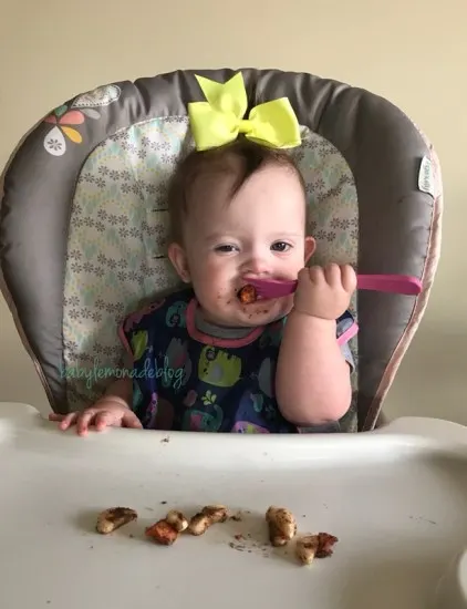 Kara in her highchair with a fork full of food and a big yellow bow in her hair
